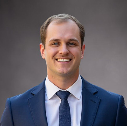 Smiling man in a navy suit and blue tie, standing against a gray backdrop.
